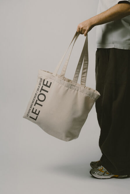 Person in white shirt holding natural canvas tote bag with Let Tote Bag Mockup text printed on it
