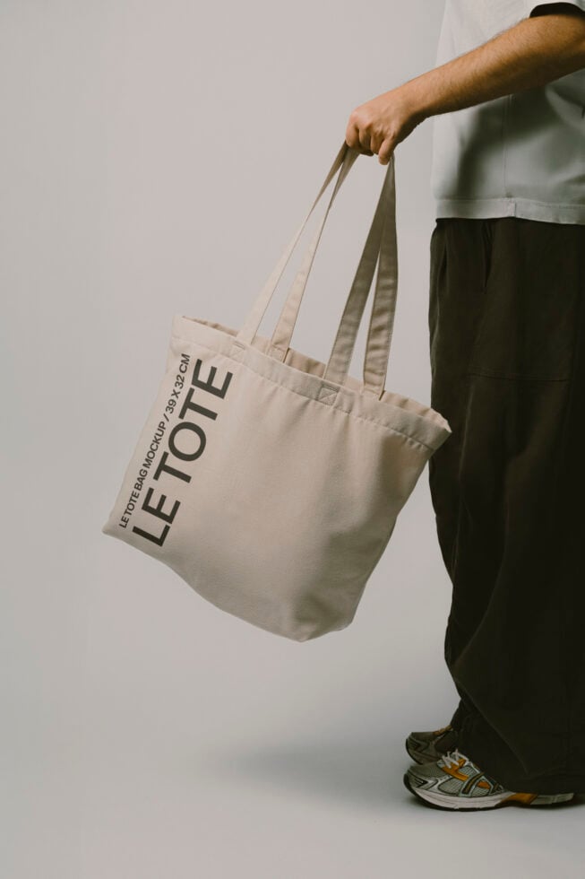 Person in white shirt holding natural canvas tote bag with Let Tote Bag Mockup text printed on it