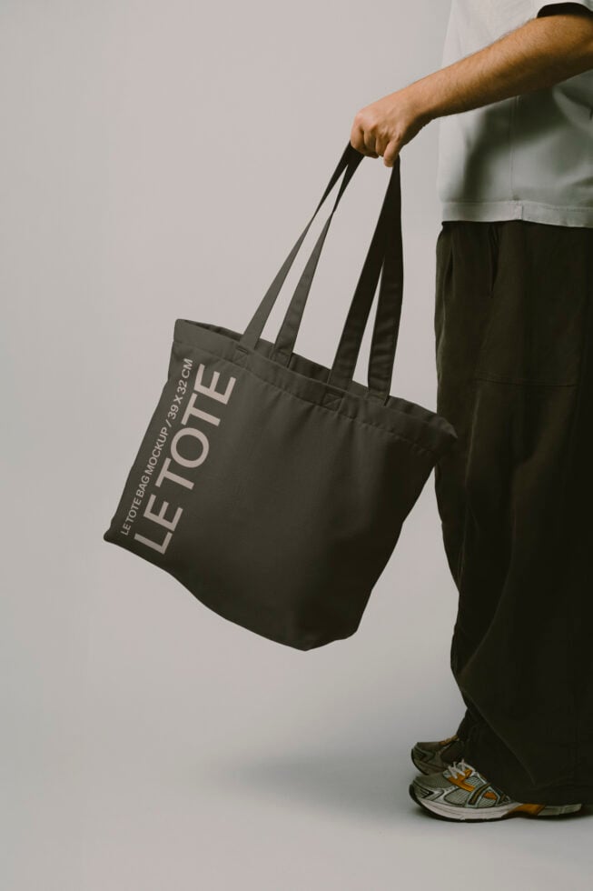 Person in white shirt holding black canvas tote bag with Let Tote Bag Mockup text printed on it