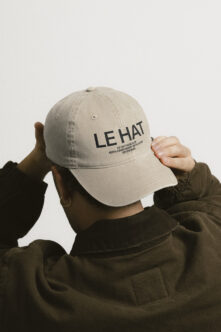 Cream colored baseball cap with LE HAT branding text worn by person in olive jacket
