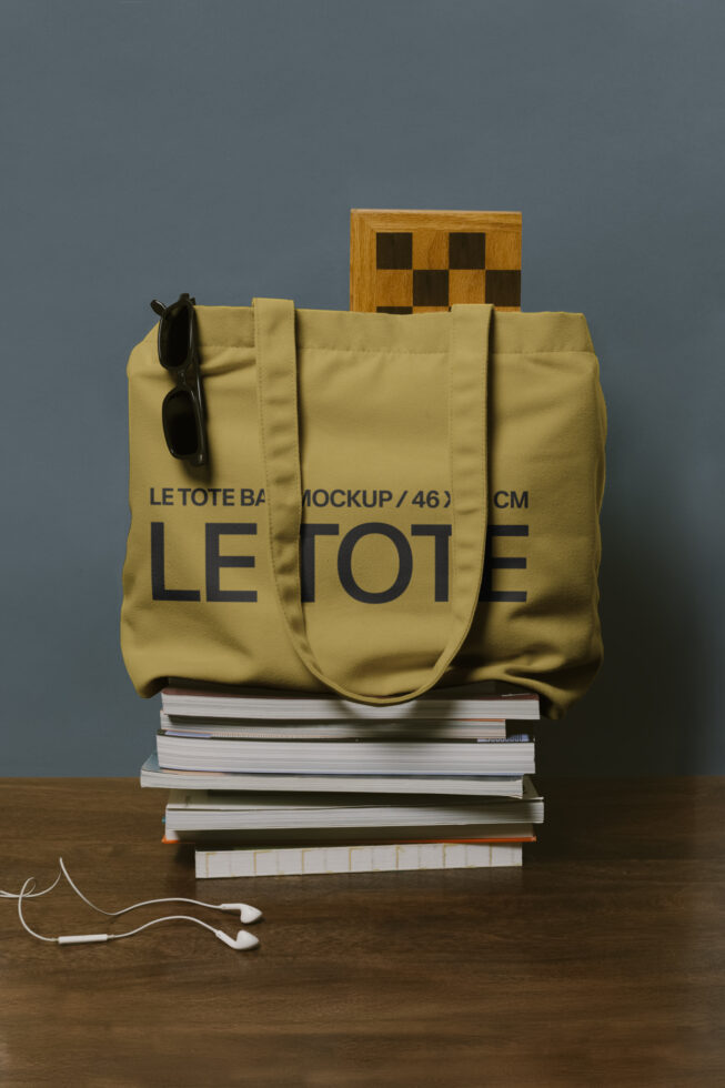 Yellow canvas tote bag with Le Tote branding stacked on books, wooden checkerboard, sunglasses attached