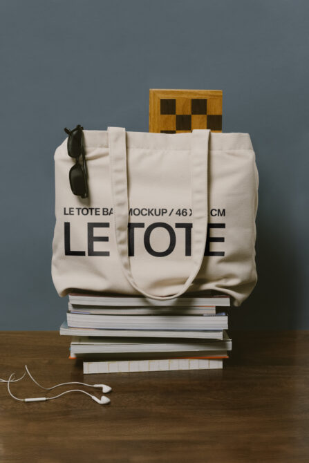 Cream canvas tote bag with Le Tote print on book stack, wooden checkerboard back, earbuds in frame