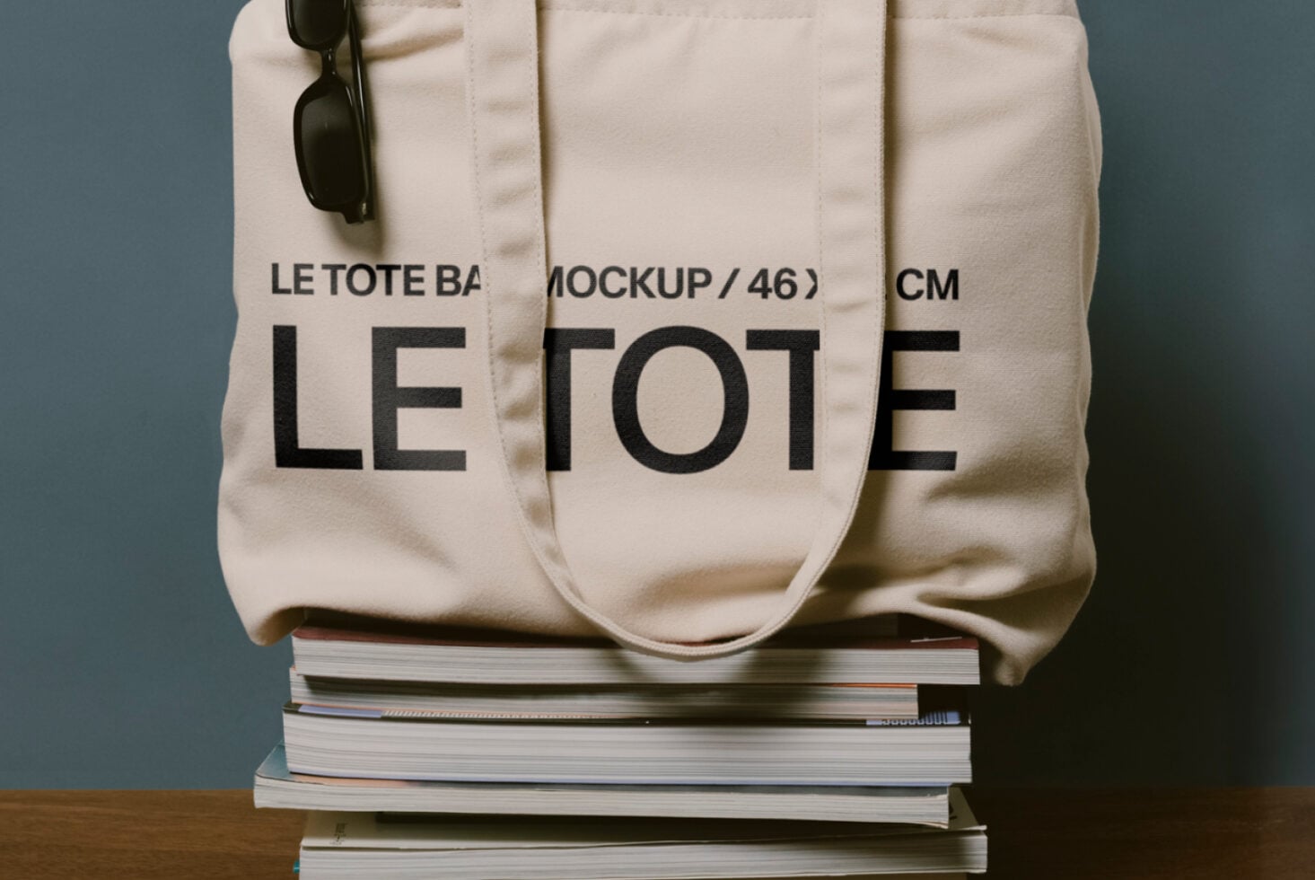 Off white canvas tote with Le Tote lettering resting on stacked books, black sunglasses visible