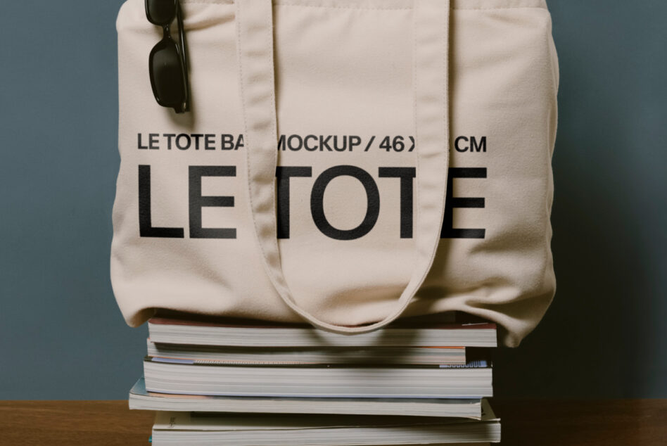 Off white canvas tote with Le Tote lettering resting on stacked books, black sunglasses visible