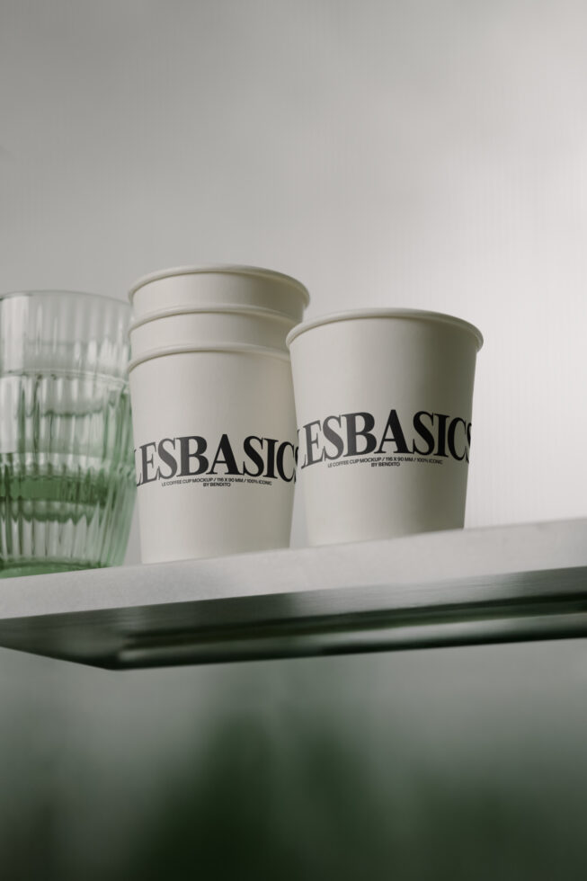 White coffee cups with Les Basics text on shelf next to green glass