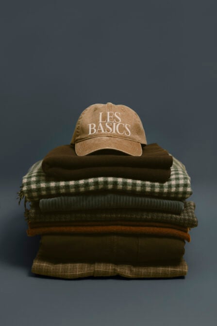 Tan baseball cap stacked on folded clothing including plaid, knit, wool textures