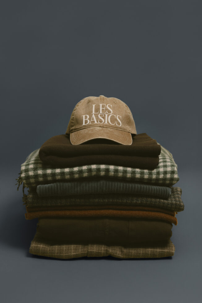 Tan baseball cap stacked on folded clothing including plaid, knit, wool textures