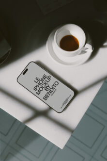iPhone with white screen mockup on white surface next to espresso cup with shadows