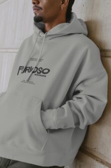 Person in light gray hoodie with dark Furioso print on chest, leaning against textured wall