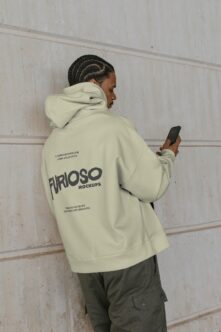 Cream hoodie back view with braided hair, holding phone against concrete wall