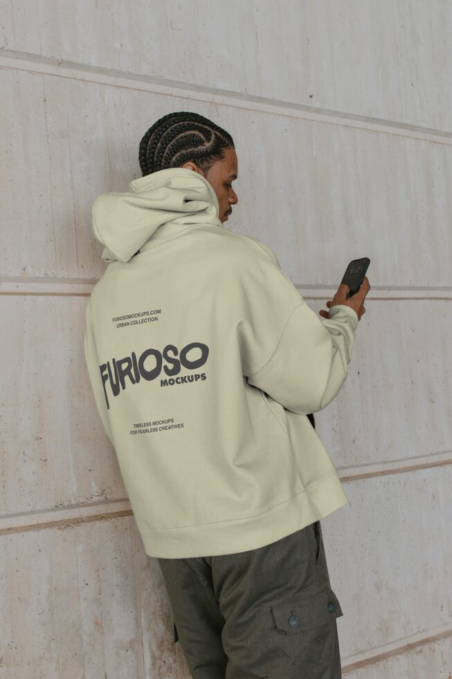 Cream hoodie back view with braided hair, holding phone against concrete wall