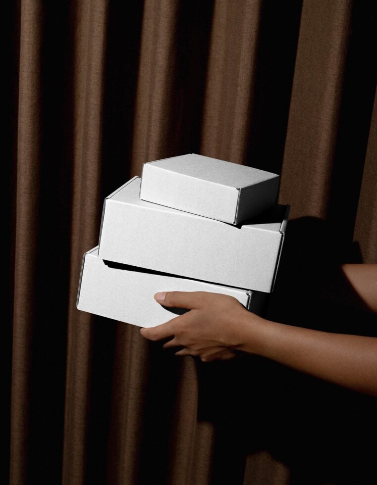 Three blank white boxes stacked in hands with brown curtain backdrop