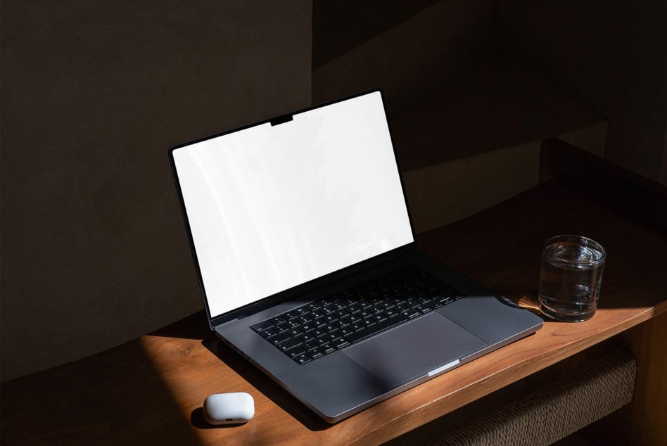 Laptop mockup with blank white screen on wooden desk, water glass nearby, dark lighting
