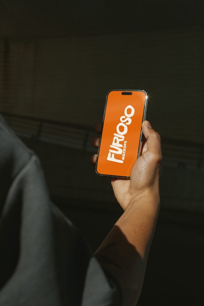 Person in dark room holding orange iPhone with Furioso Mockups logo centered