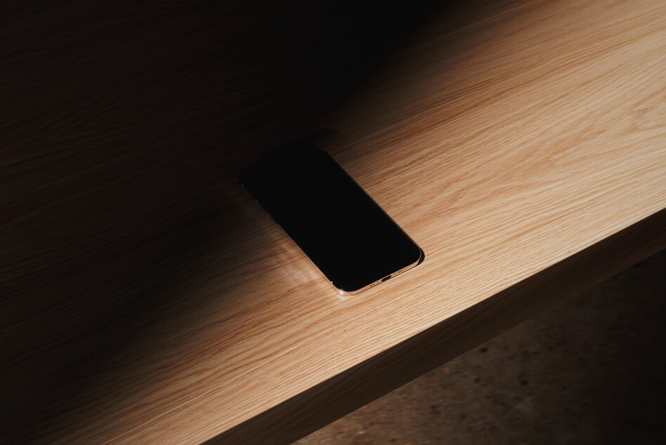 iPhone with blank black screen resting on light wood surface with soft shadows