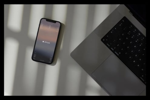 iPhone mockup displaying gradient akoya app interface with laptop keyboard on minimal desk