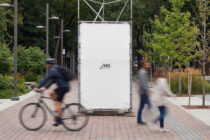 Blank white billboard mockup in tree-lined plaza with moving people