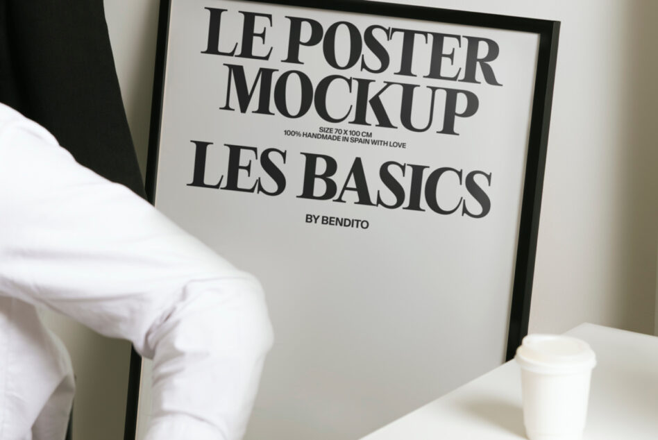 White poster in black frame showing Les Basics branding, styled with coffee on desk