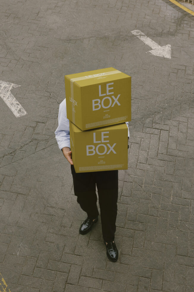 Person holding stacked yellow boxes labeled Le Box on urban pavement