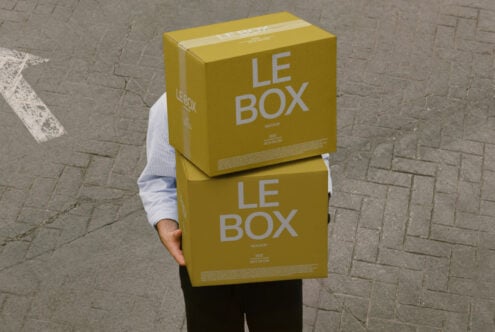 Person carrying two yellow cardboard boxes labeled LE BOX outdoors