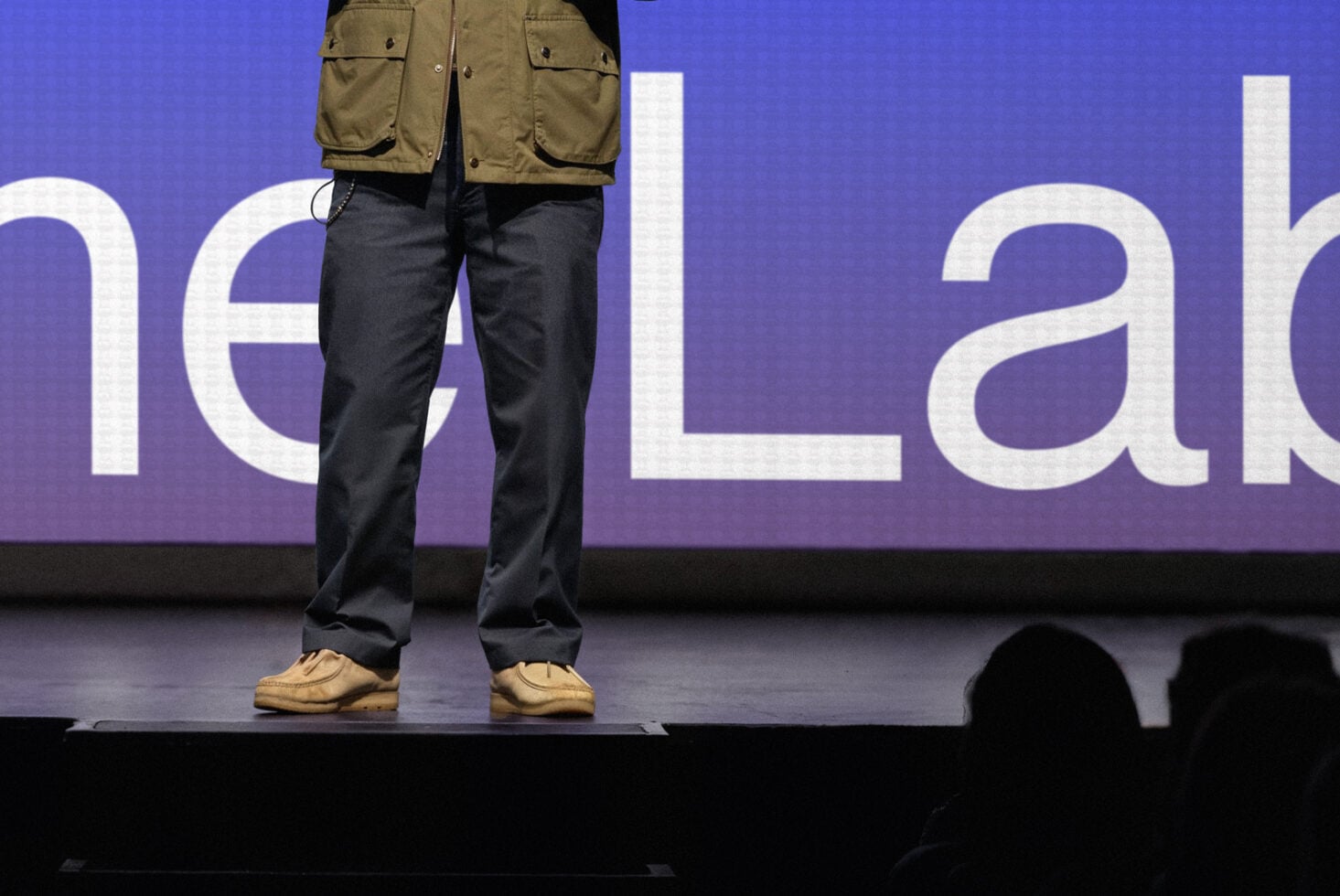 Close-up of legs on stage with khaki jacket, dark jeans, tan shoes