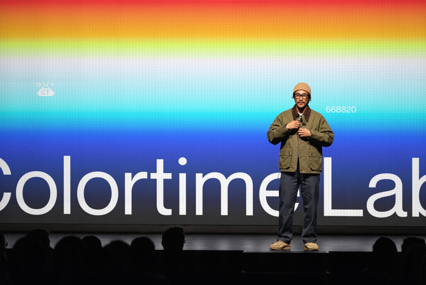 Speaker on stage with colorful gradient backdrop, audience silhouettes below