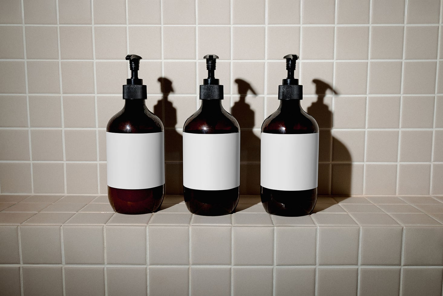 Three dark glass pump bottles with white label bands lined up on cream tile shelf