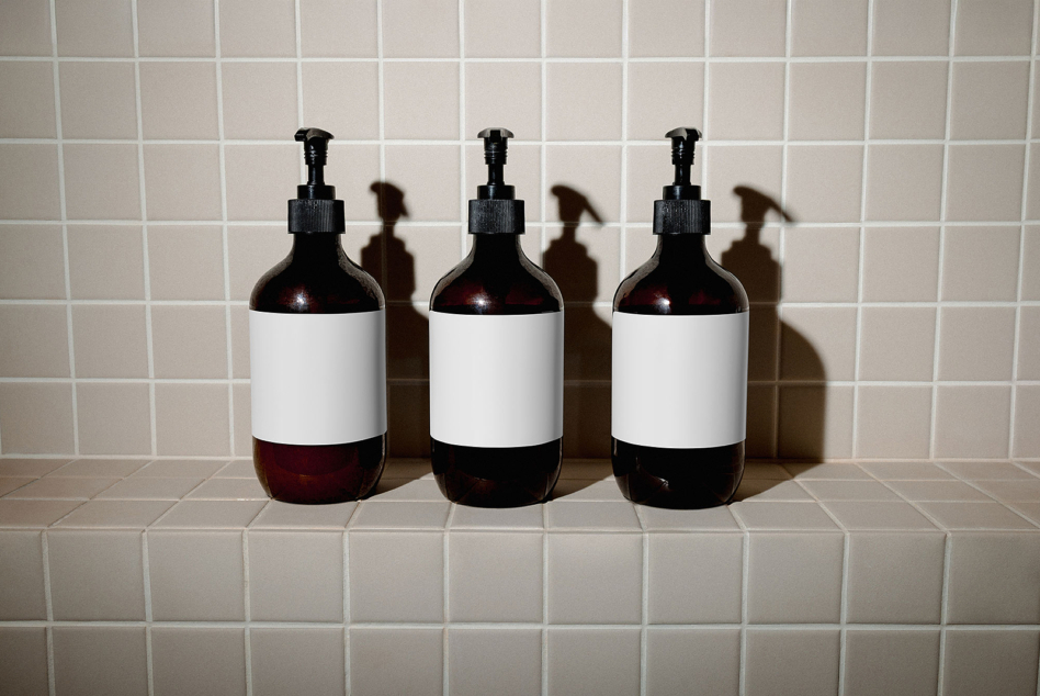 Three dark glass pump bottles with white label bands lined up on cream tile shelf