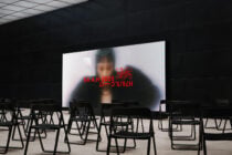Large display screen showing blurred figure with red glitch text overlay in empty auditorium