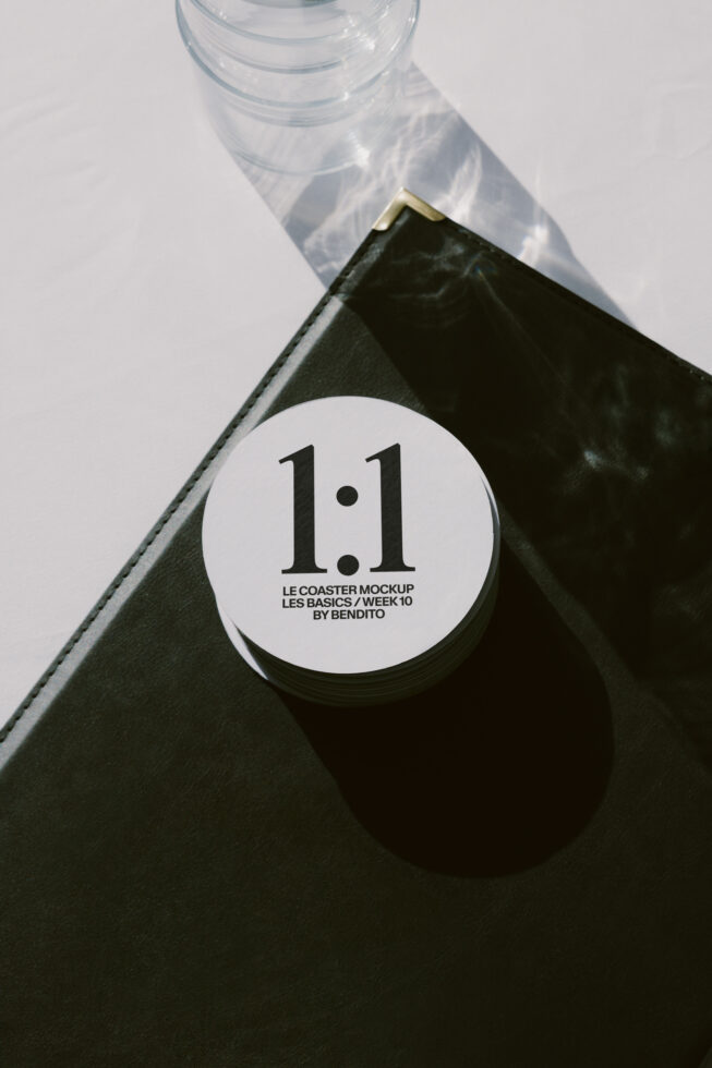 White circular coaster mockup on black leather notebook with water glass