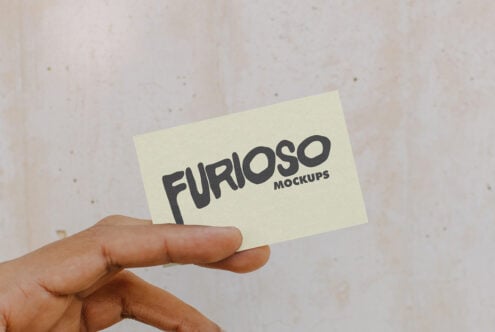 Fingers gripping light cream business card with dark Furioso Mockups logo on beige surface