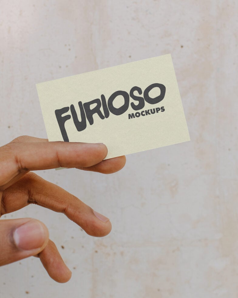 Hand holding light yellow business card with dark Furioso Mockups logo on neutral surface