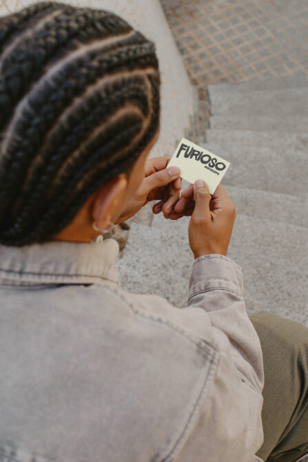 Hands presenting cream-colored square business card with Furioso Mockups text outdoors