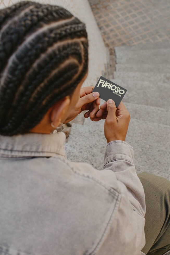 Person with braided hair holding dark square business card showing Furioso Mockups branding
