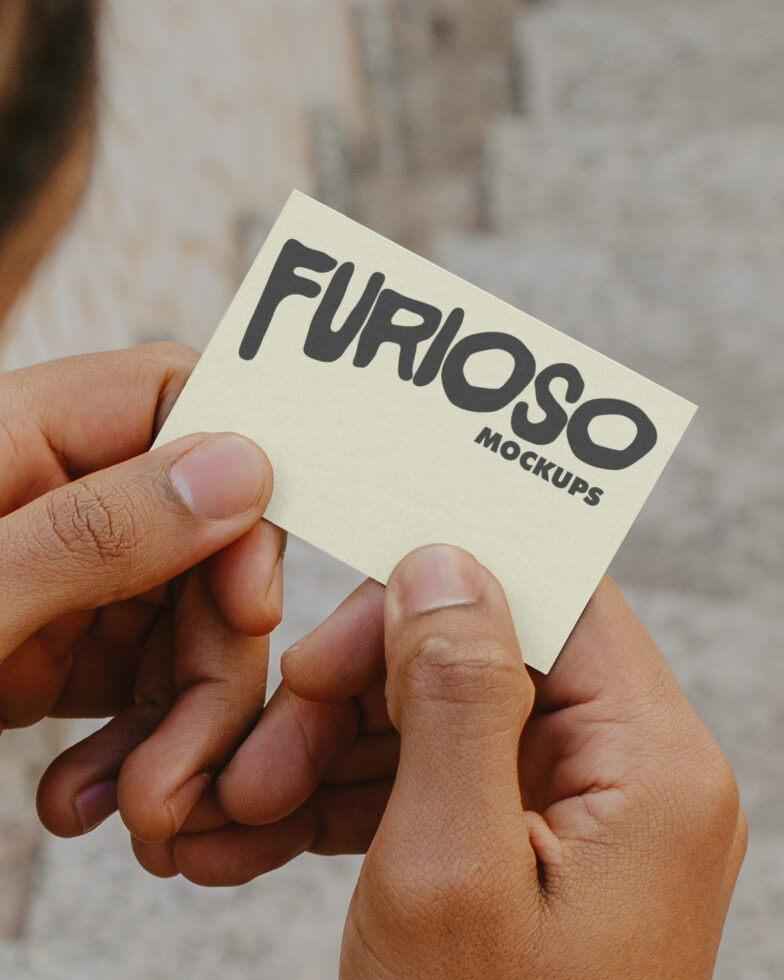 Hands holding small cream square business card showing Furioso Mockups logo against blurred background