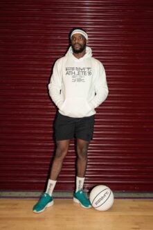 Guy in cream hoodie with Athlete branding standing against red corrugated wall