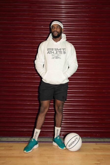 Guy in cream hoodie with Athlete branding standing against red corrugated wall