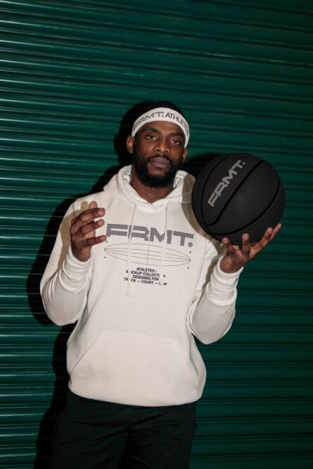 Guy in cream hoodie with teal metal backdrop holding black basketball