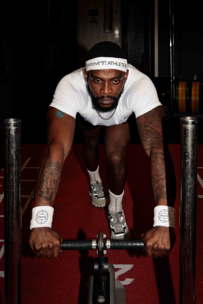 Athlete in white FRMT tee with headband leaning over dumbbell on red mat, gym setting