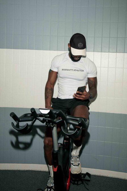 Man on stationary bike wearing white FRMT Athlete tee, holding phone, tiled wall backdrop
