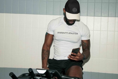 Man seated on bike in white FRMT Athlete tee, black cap visor, holding phone