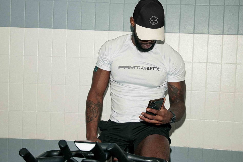 Man seated on bike in white FRMT Athlete tee, black cap visor, holding phone