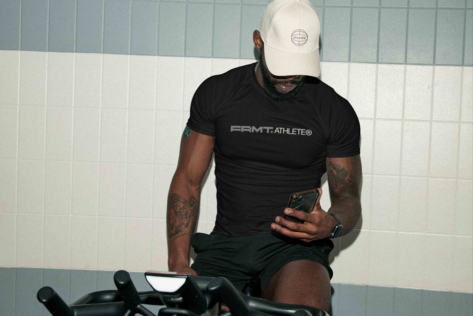 Man on stationary bike in black FRMT Athlete shirt, white cap, checking phone