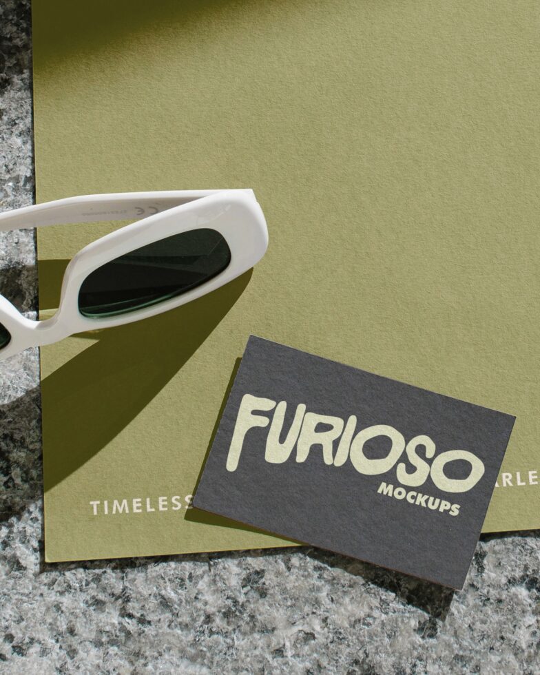 Dark business card sits on olive tone notepad next to white oval sunglasses