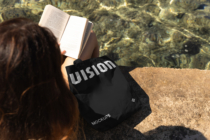 Woman reading open book by water wearing black canvas tote bag with white text
