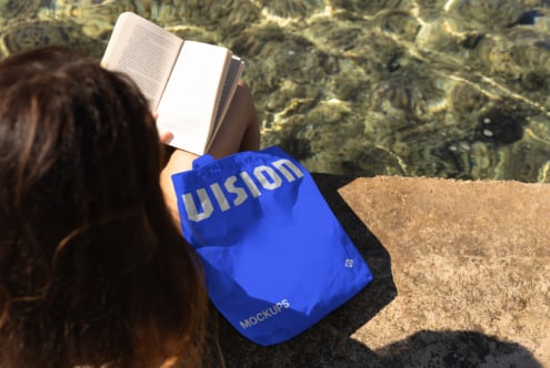 Woman reading open book by water wearing blue canvas tote bag with white text