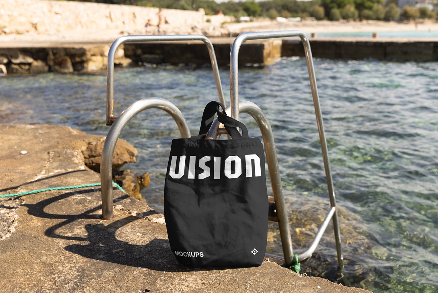 Black canvas tote bag hanging on metal railing by ocean with rope visible