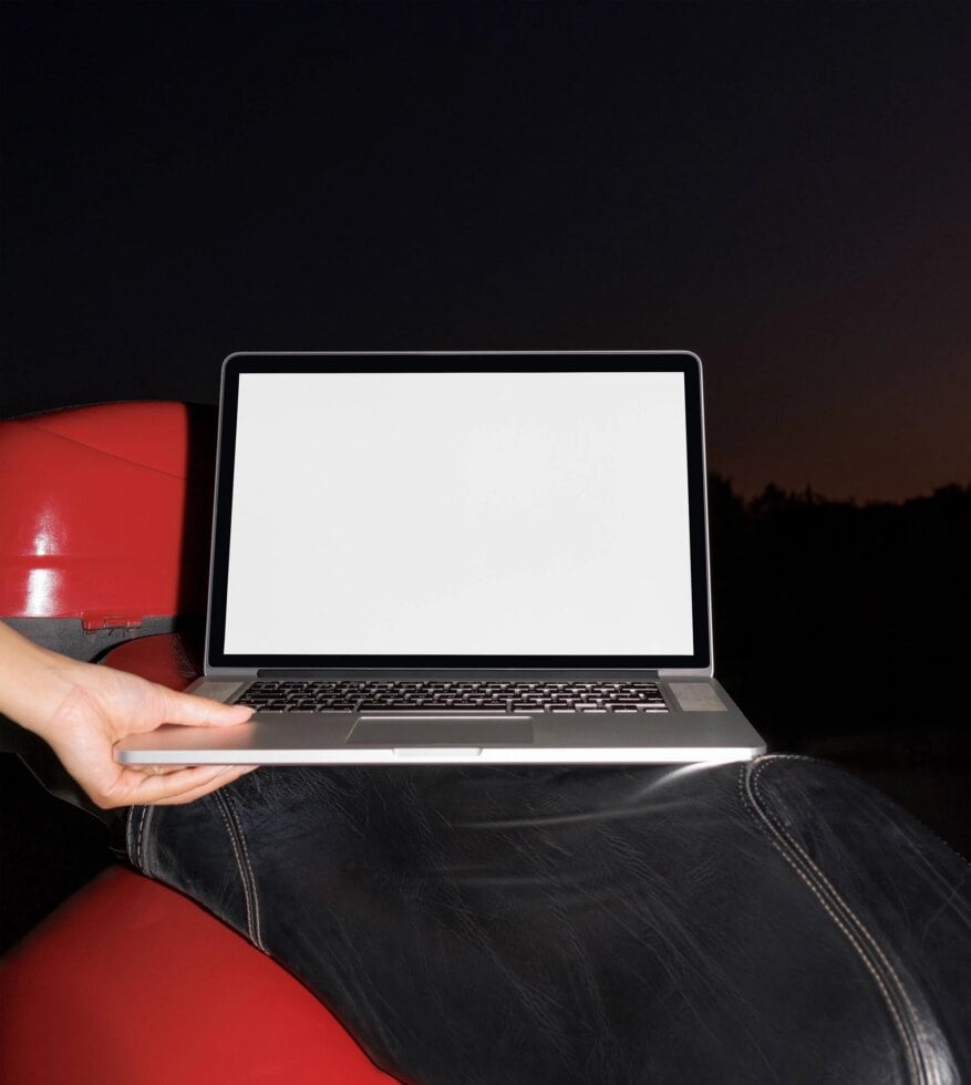 Laptop mockup on a motorcycle seat, held by hand, blank screen for design presentation, nighttime background, ideal for showcasing digital assets