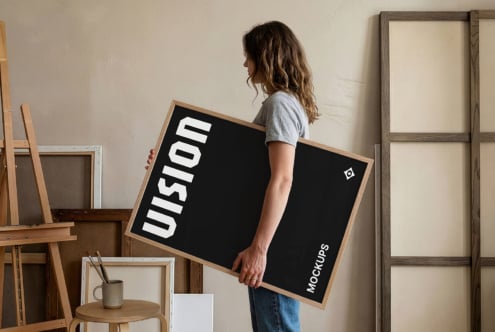 Woman in gray tee holding wooden-framed black mockup poster in studio setting