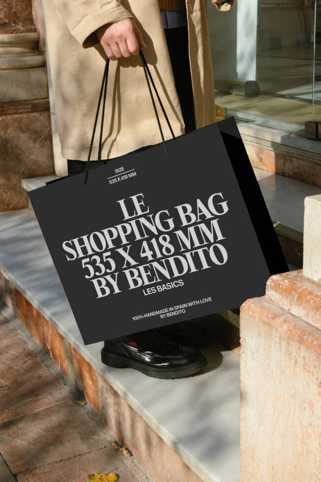 Black shopping bag with white serif typography held by person in beige coat